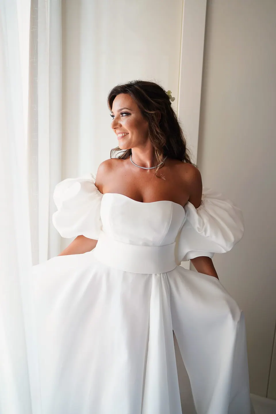 Bride standing near a window in her voluminous gown, natural light creating a soft glow.