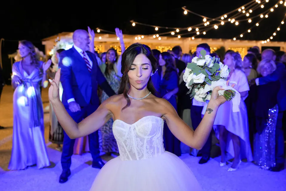 Bride raising her bouquet triumphantly beneath twinkling lights, surrounded by friends and celebration.