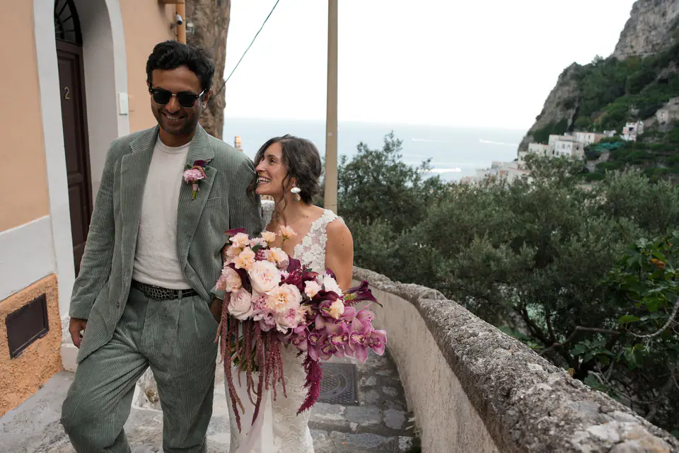 Groom laughing with the bride on a stone staircase, the sea behind them.