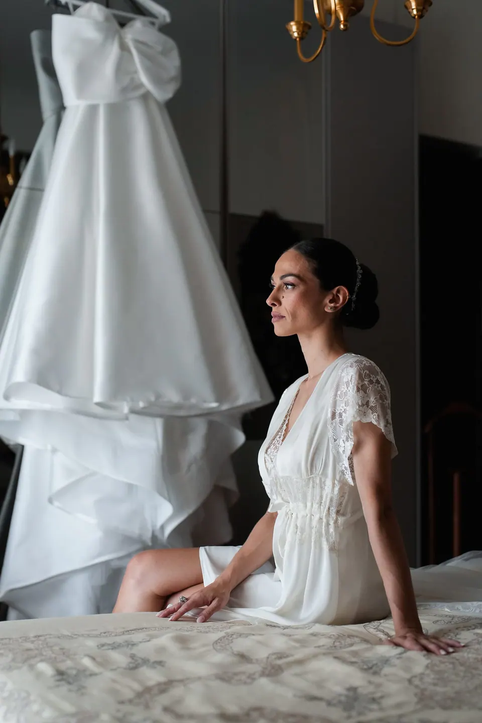 Sposa in vestaglia di pizzo seduta sul letto accanto all'abito appeso.