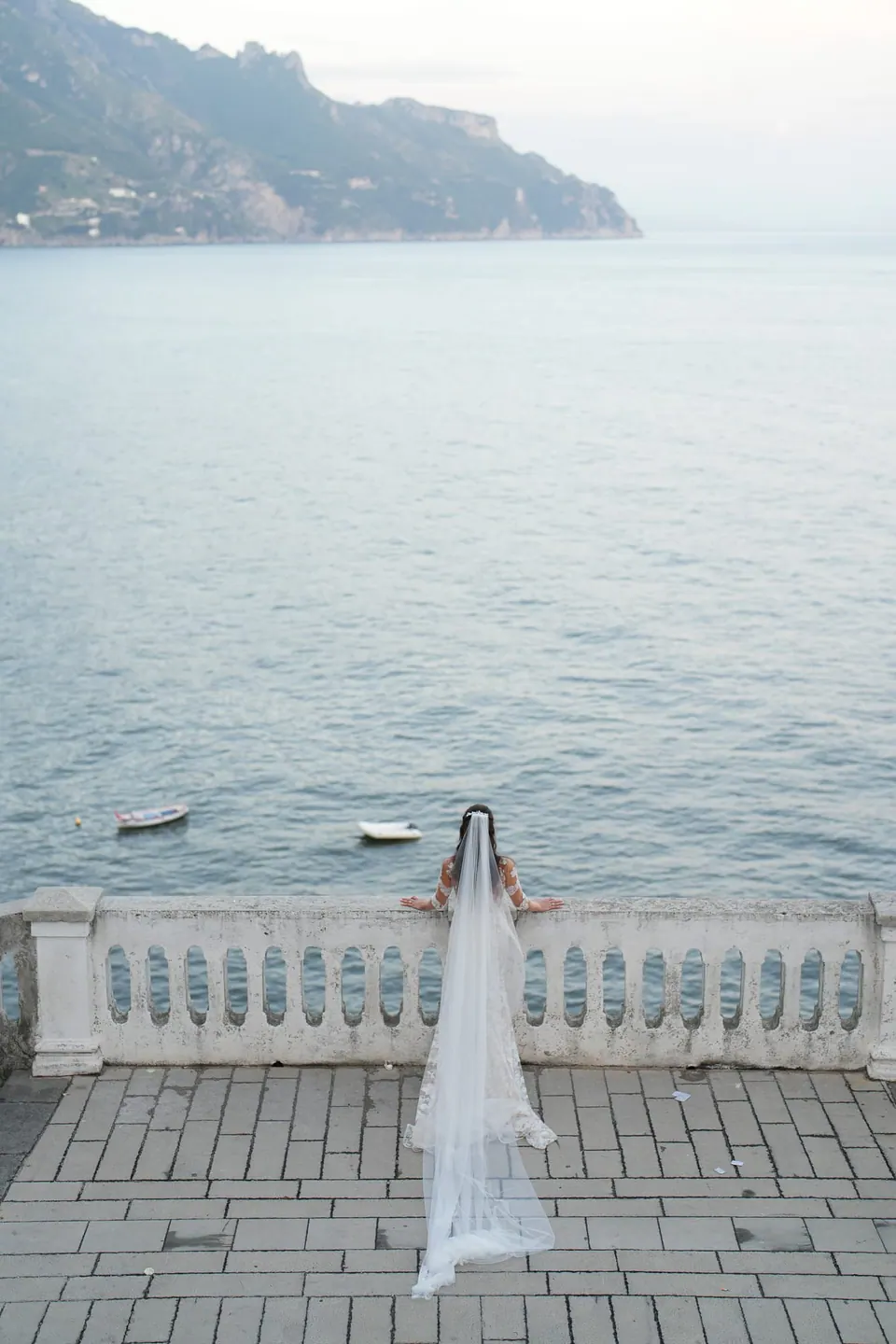 Die Braut von hinten an einer Balustrade über dem Meer, ihr Schleier hängt herab.