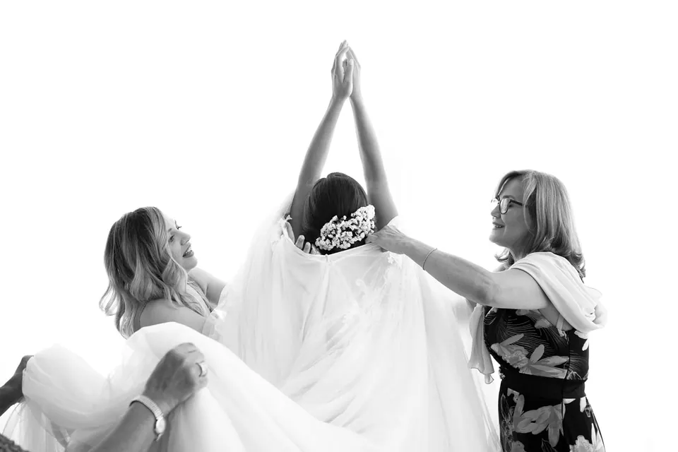 Foto in bianco e nero della sposa che alza le braccia mentre le parenti sollevano abito e velo.