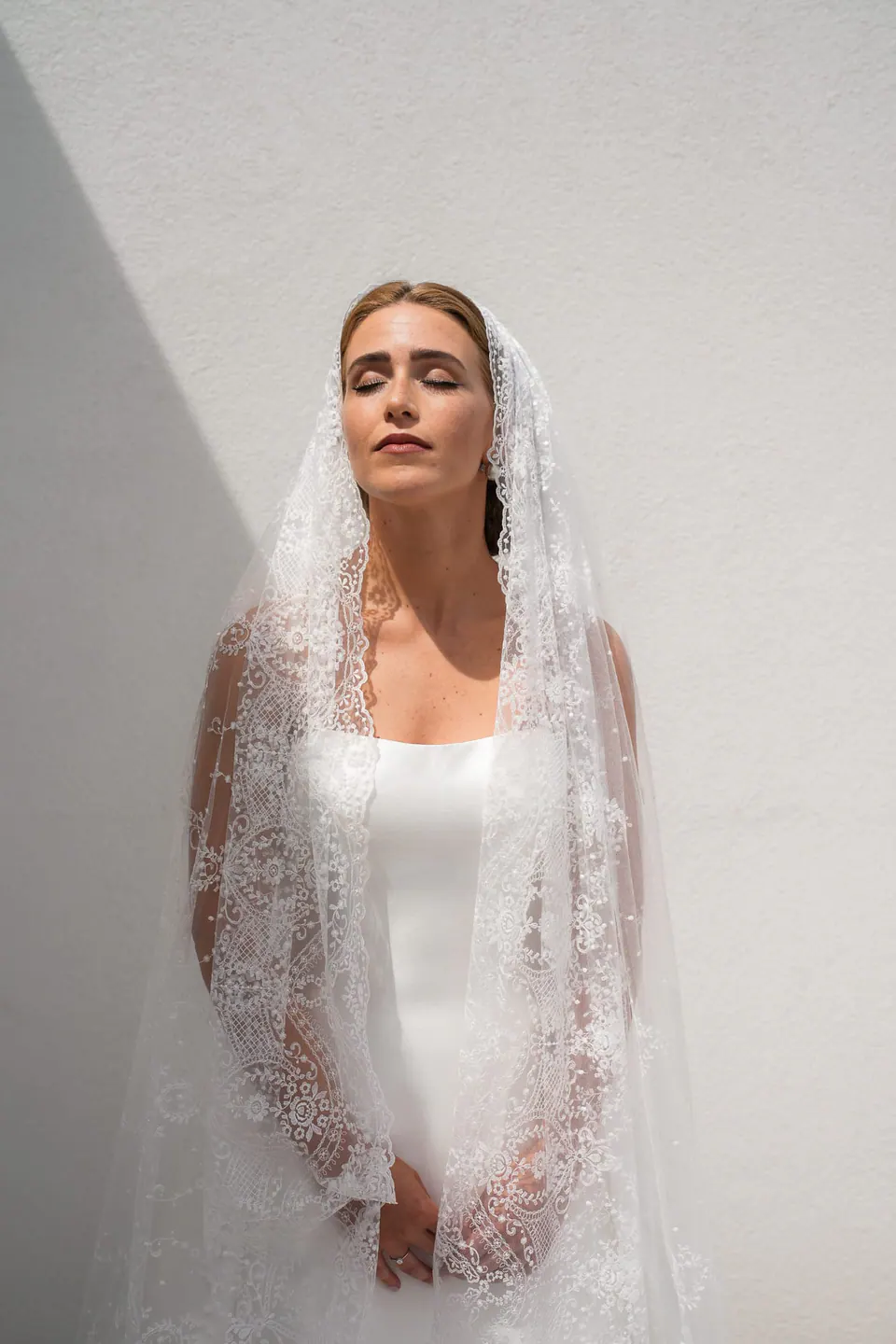 Die Braut mit geschlossenen Augen unter ihrem Spitzenschleier vor einer weißen Wand.