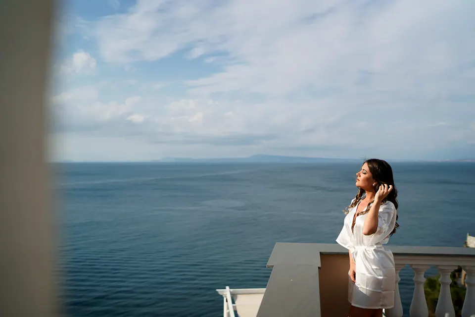 Bride on the terrace at the edge of the frame, the open sea beyond.