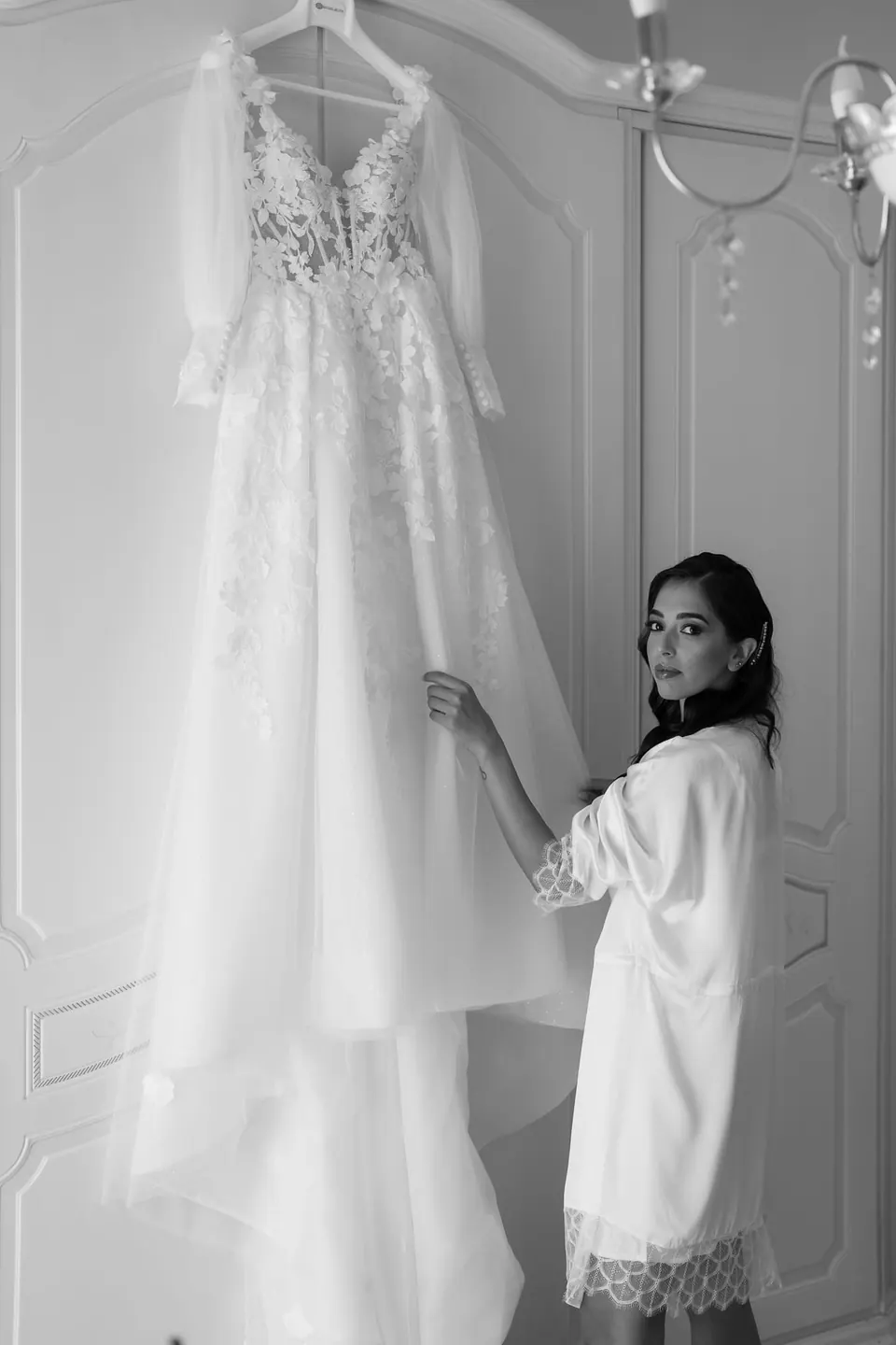 Black and white photo of the bride in her robe beside the gown hanging on the wardrobe.