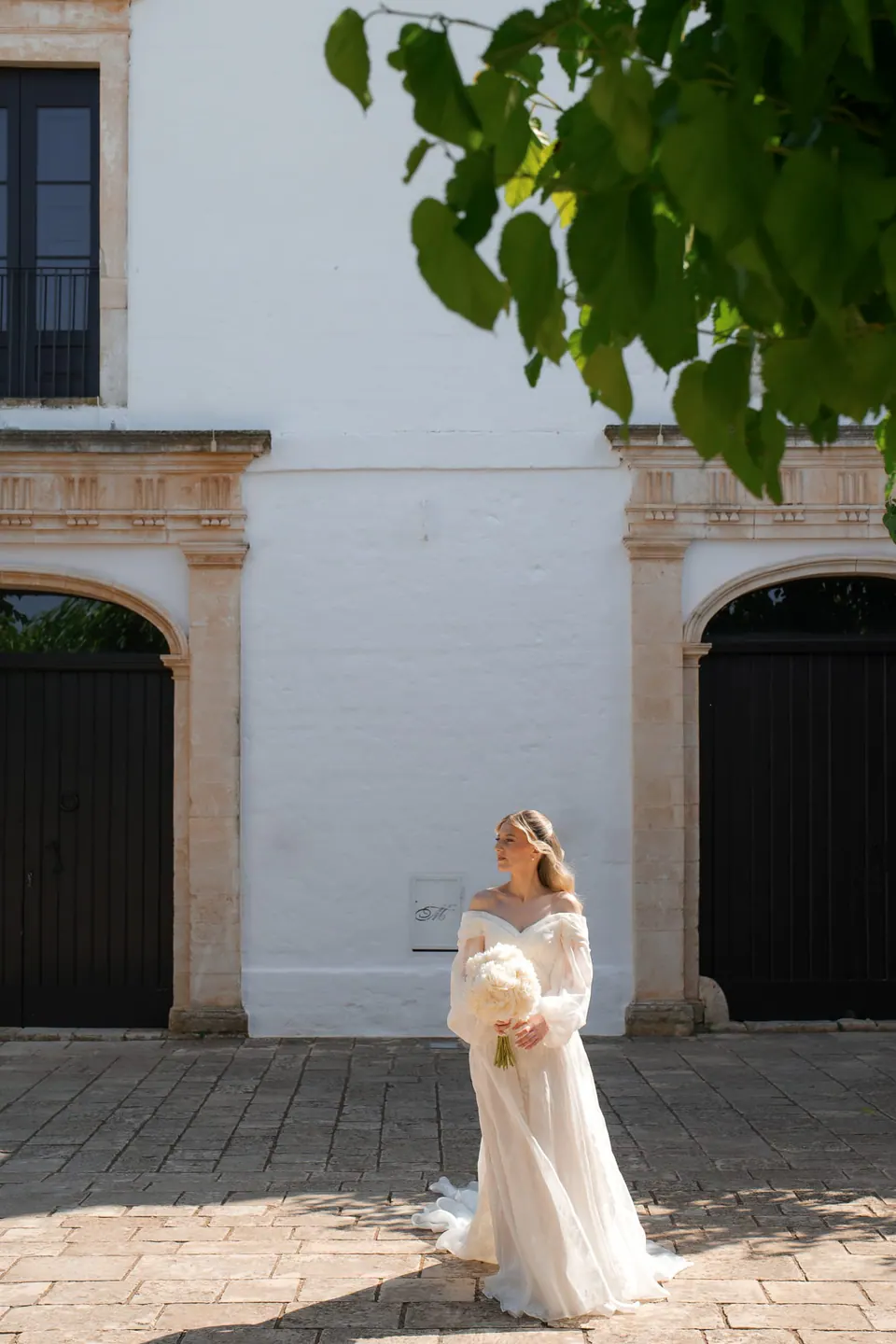 La novia pasa junto a muros blancos bajo los árboles.