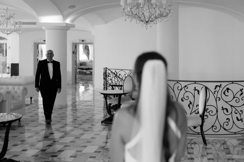 Black and white photo of the groom waiting in the hall as the bride approaches from behind.