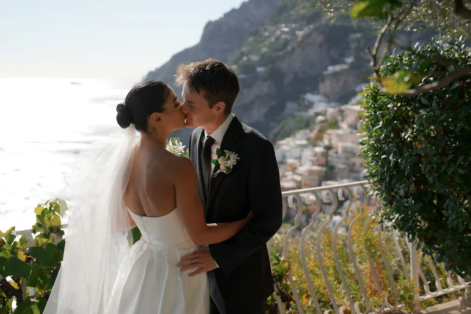 Bride and groom kissing among the greenery, her veil in the light.