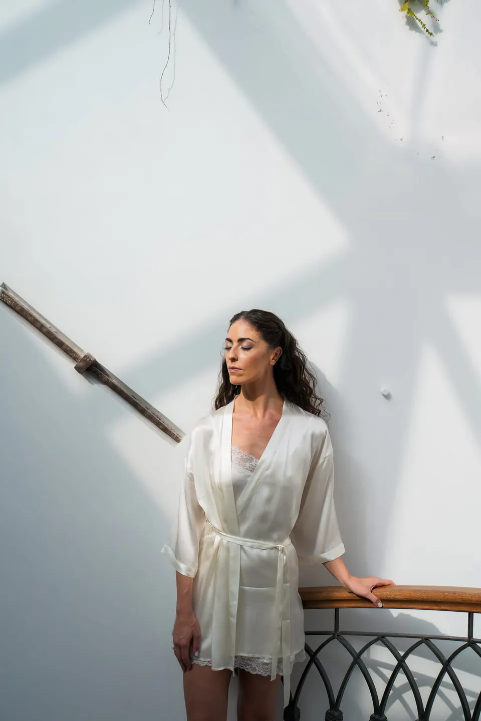 Bride in a satin robe on the stair landing, soft light on the white walls.