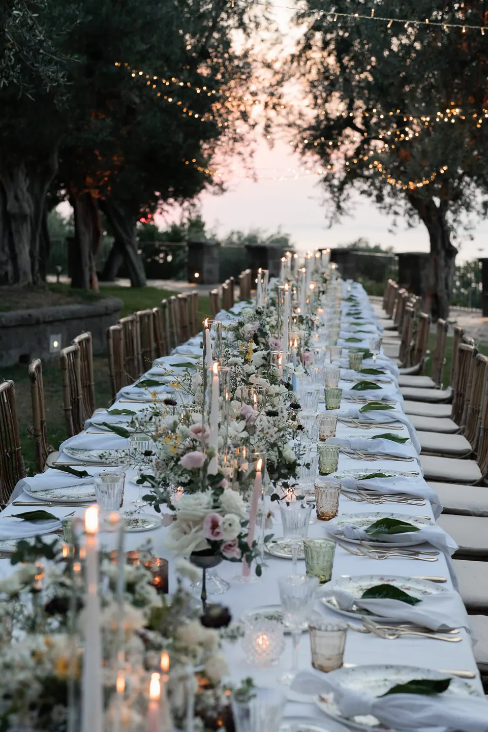 Long dinner table surrounded by olive trees and Mediterranean greenery.