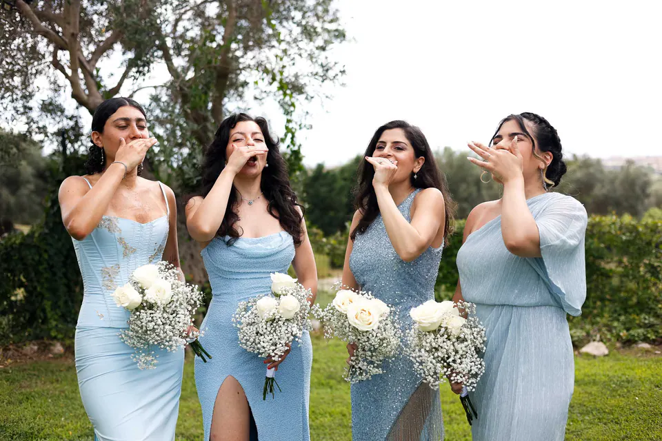 Quatre demoiselles d’honneur en bleu clair boivent un shot ensemble, bouquets de roses blanches à la main.