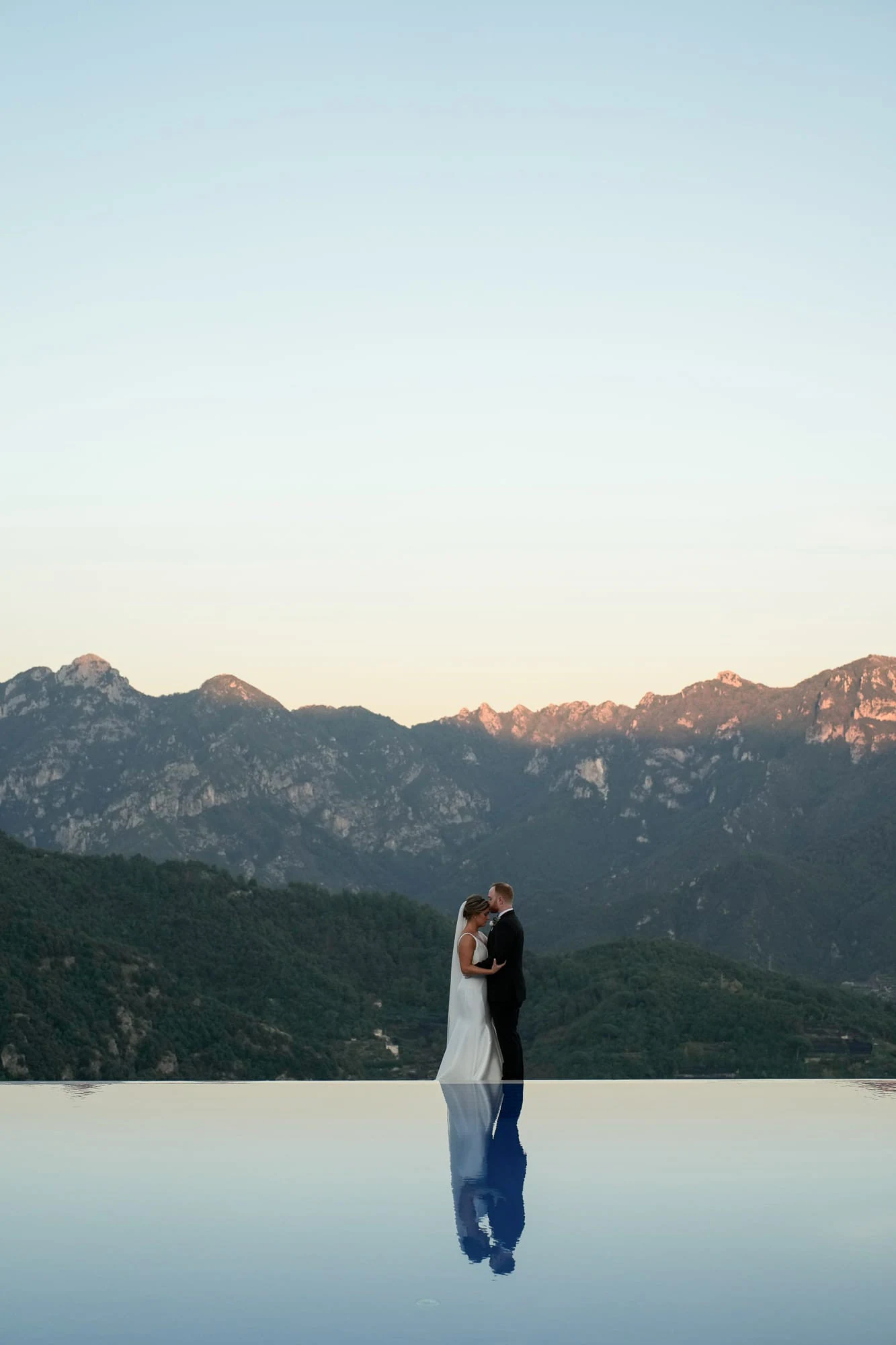 Bride and groom at the edge of the infinity pool of Belmond Hotel Caruso, the mountains reflected in the water.