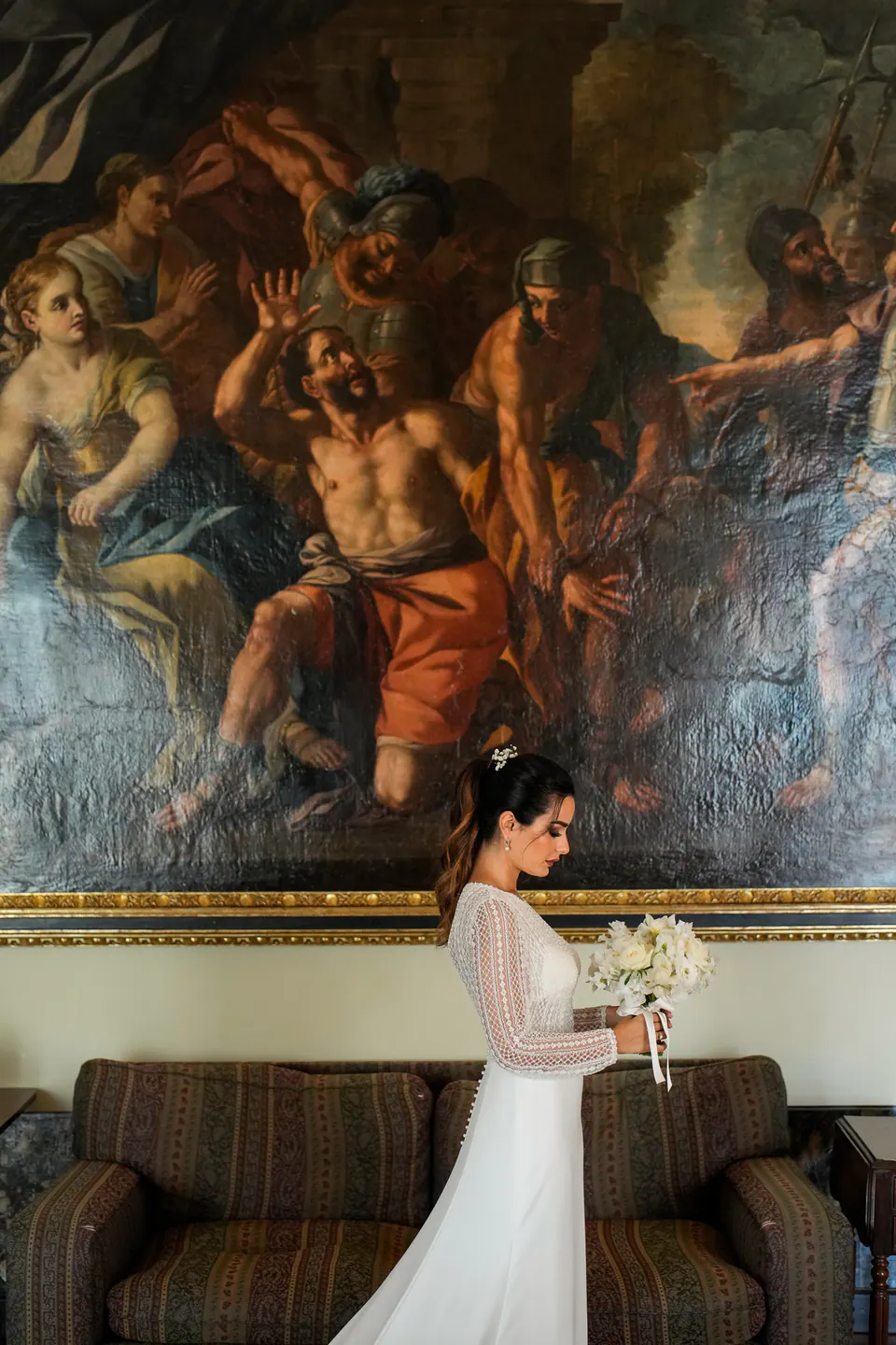 Bride adjusting her dress in front of a baroque painting, soft light illuminating her face.