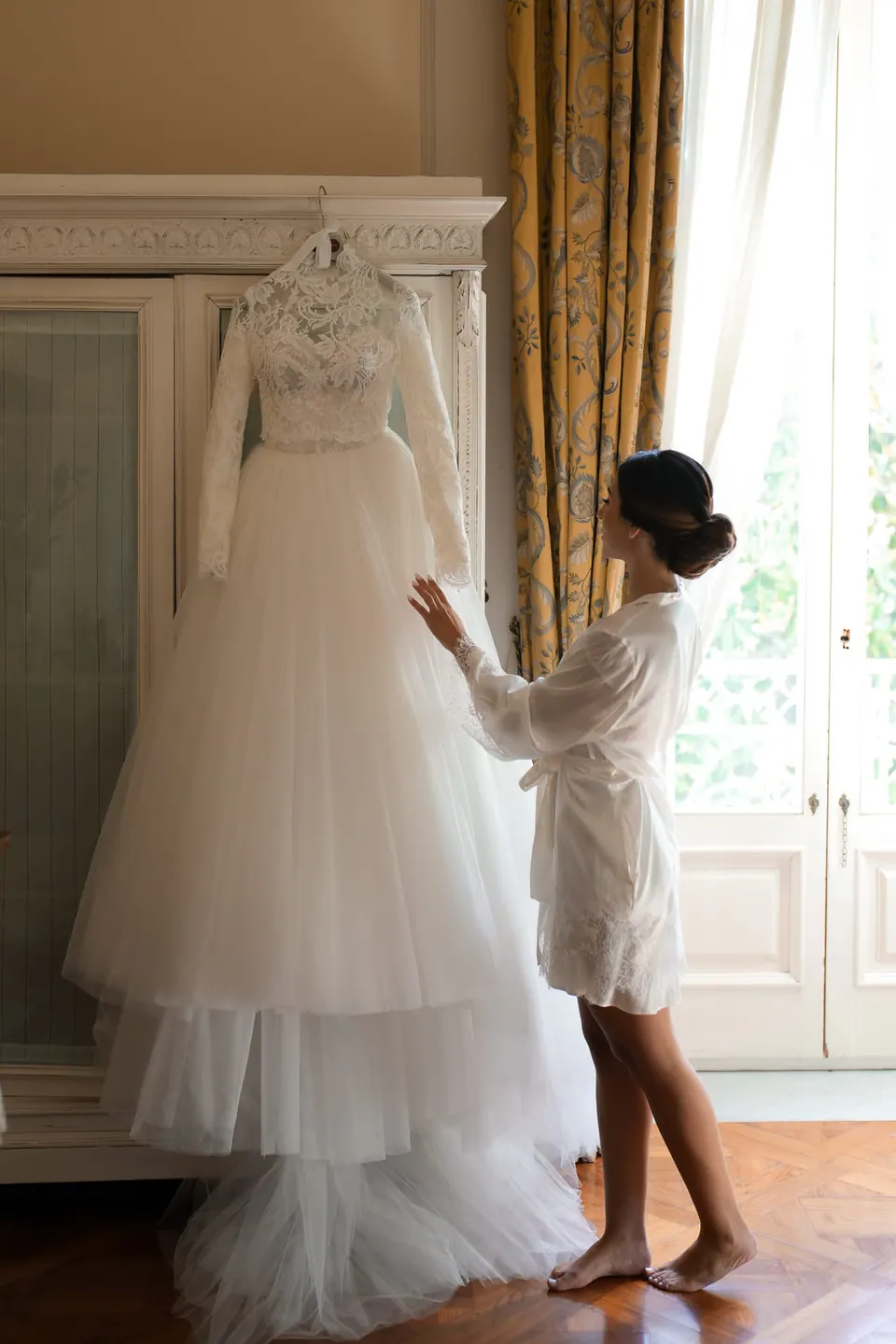Bride touching her wedding dress hanging by the window
