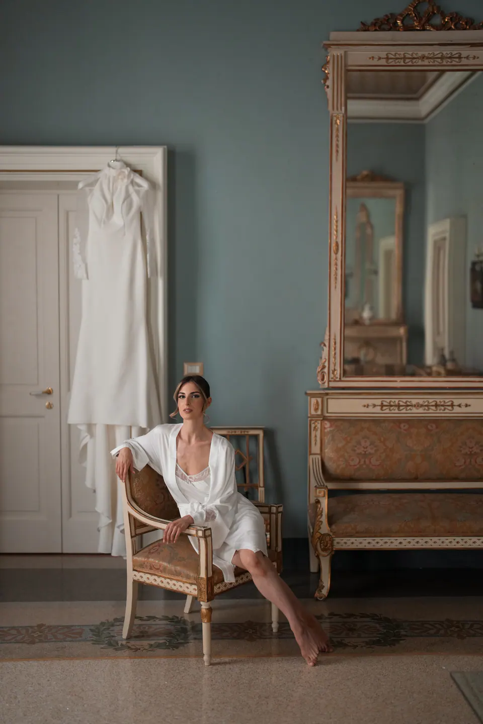 La sposa in vestaglia bianca seduta su una sedia dorata, con l'abito appeso alla porta alle sue spalle.