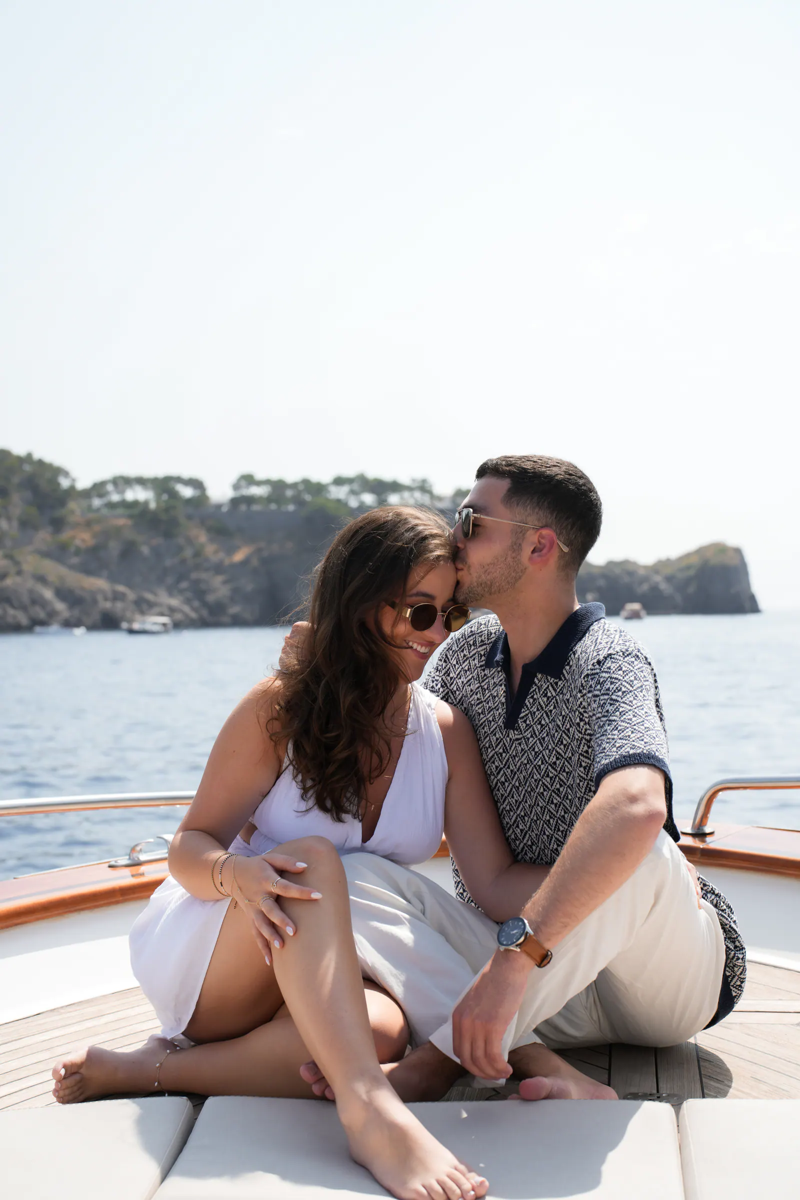 Couple sitting close on the bow of the boat, a rocky islet behind them.
