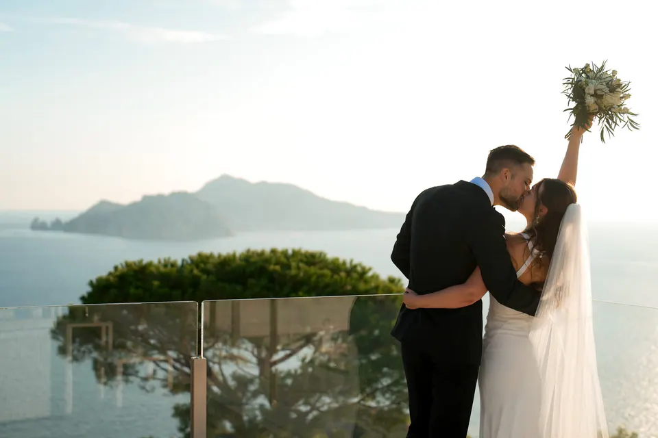 Bride and groom kissing as she raises her bouquet, Capri across the sea behind them.