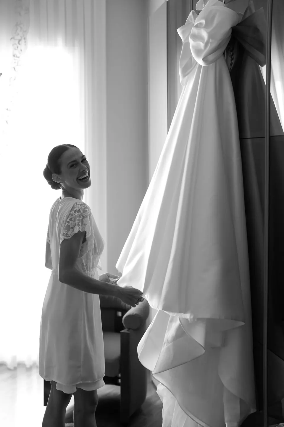 Foto in bianco e nero della sposa in vestaglia vicino alla finestra accanto all'abito appeso.