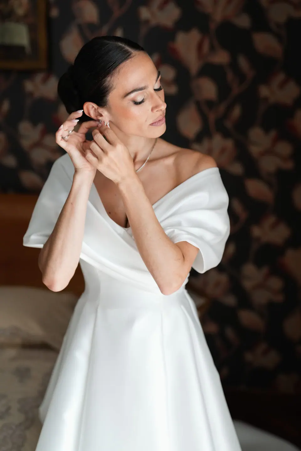 La sposa indossa gli orecchini davanti alla carta da parati scura a fiori.