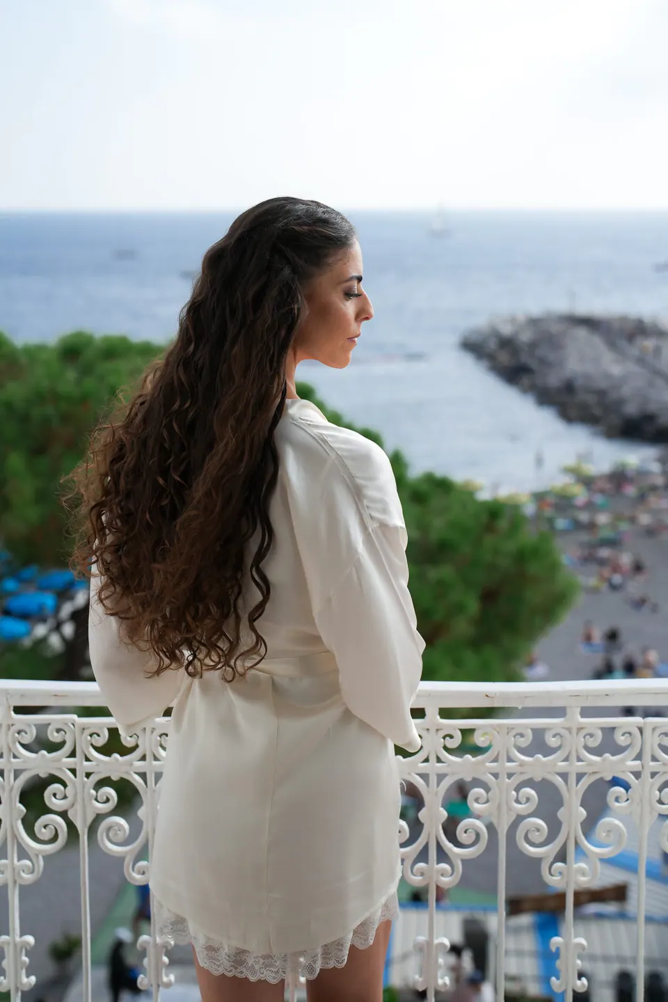 Bride in her robe on the balcony seen from behind, looking at the sea.