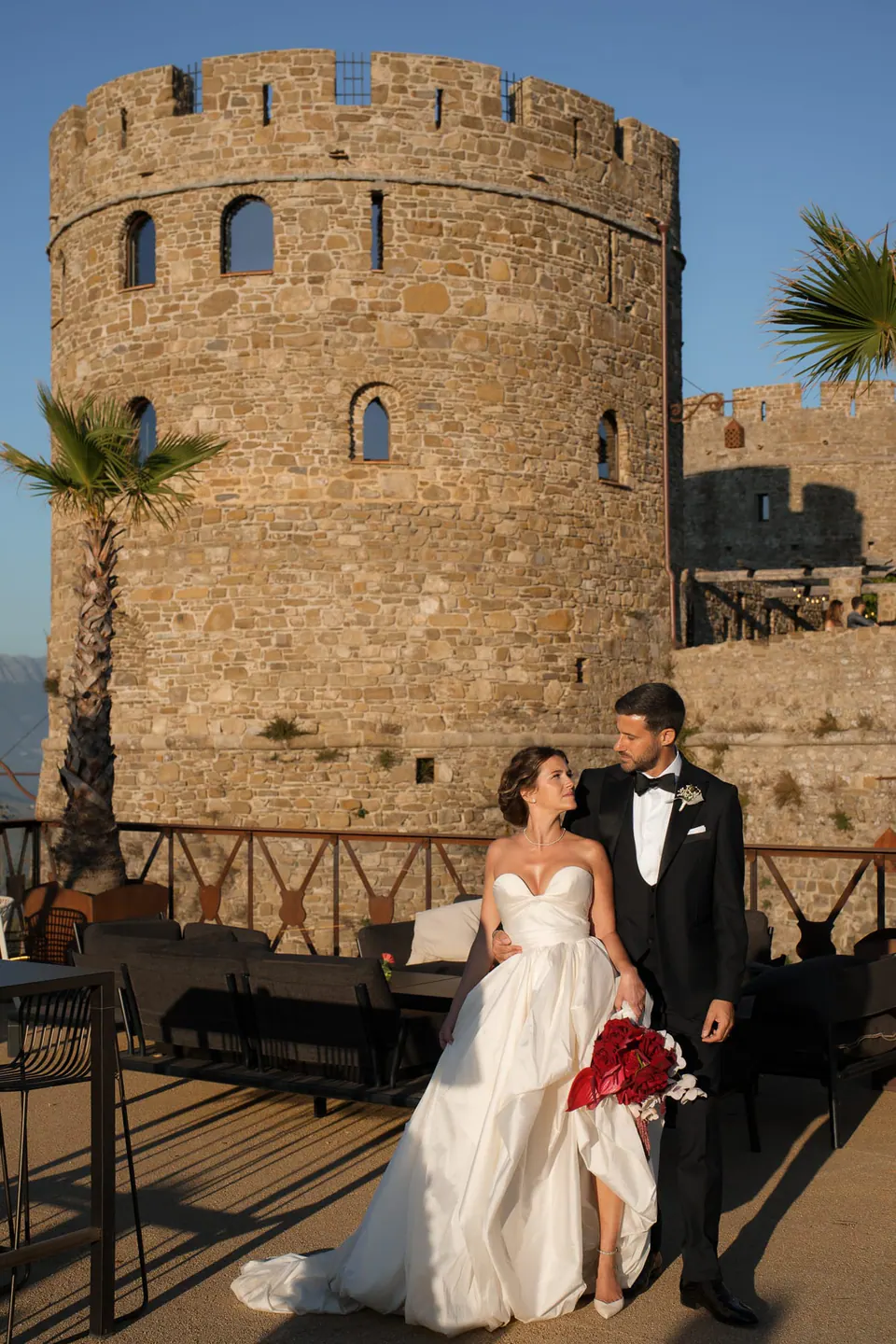 Sposi sulla terrazza davanti alla torre rotonda medievale, palme alle spalle.