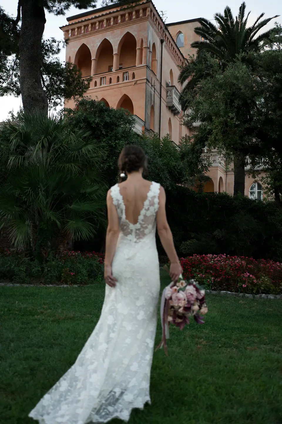 Bride walking across the lawn of a villa garden, seen from behind.