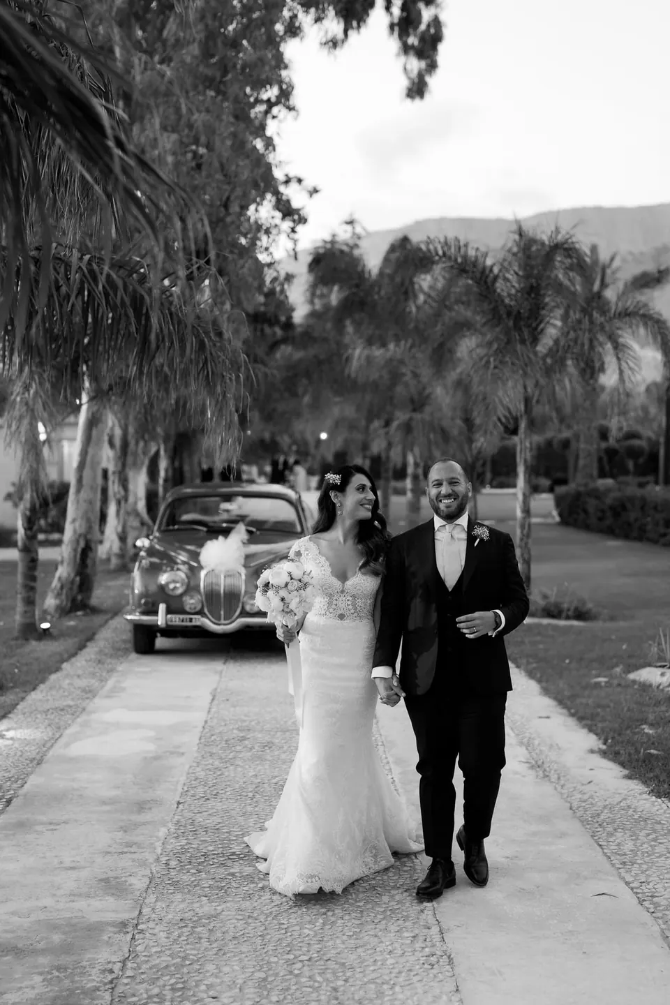 Black and white photo of bride and groom walking under palms with a vintage car behind