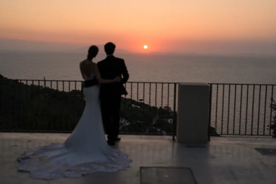 Couple from behind at a railing, watching the sunset over the sea.