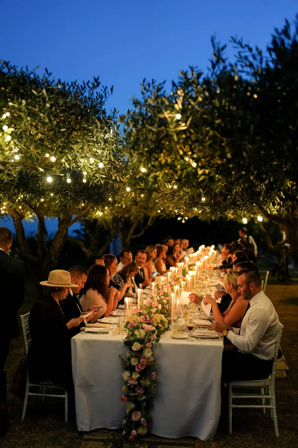 Long wedding dinner table under olive trees strung with lights at dusk, Belmond Hotel Caruso