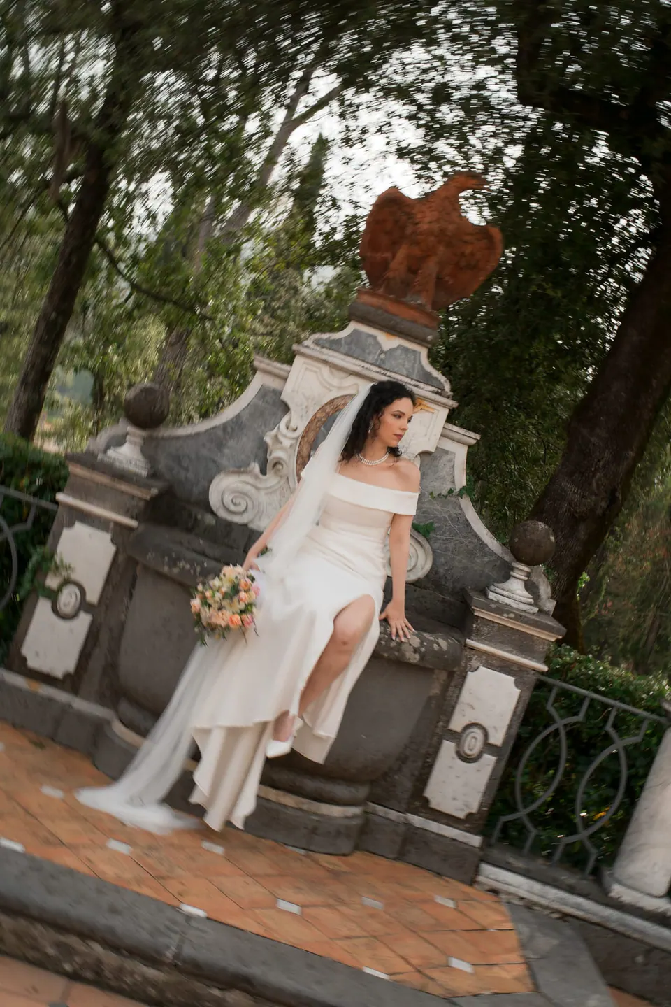 La novia sentada en el borde de una fuente barroca en los jardines de la villa, con vestido de hombros descubiertos y ramo.