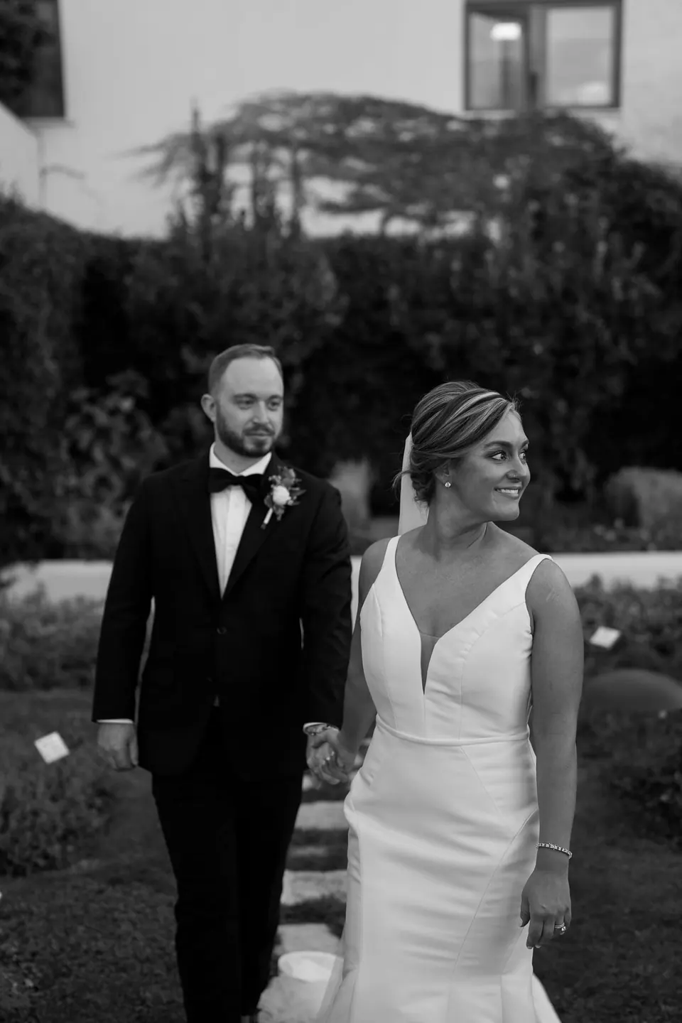 Black and white photo of bride and groom walking through the garden in the evening