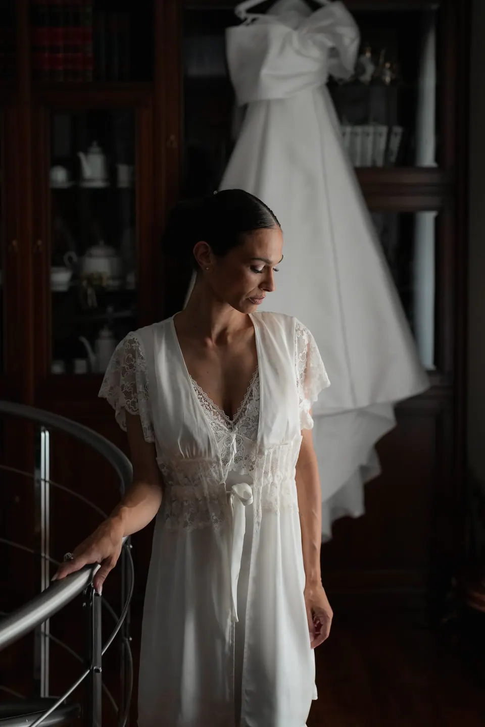 Bride in a lace robe at the bottom of the spiral stairs, the gown hanging above.