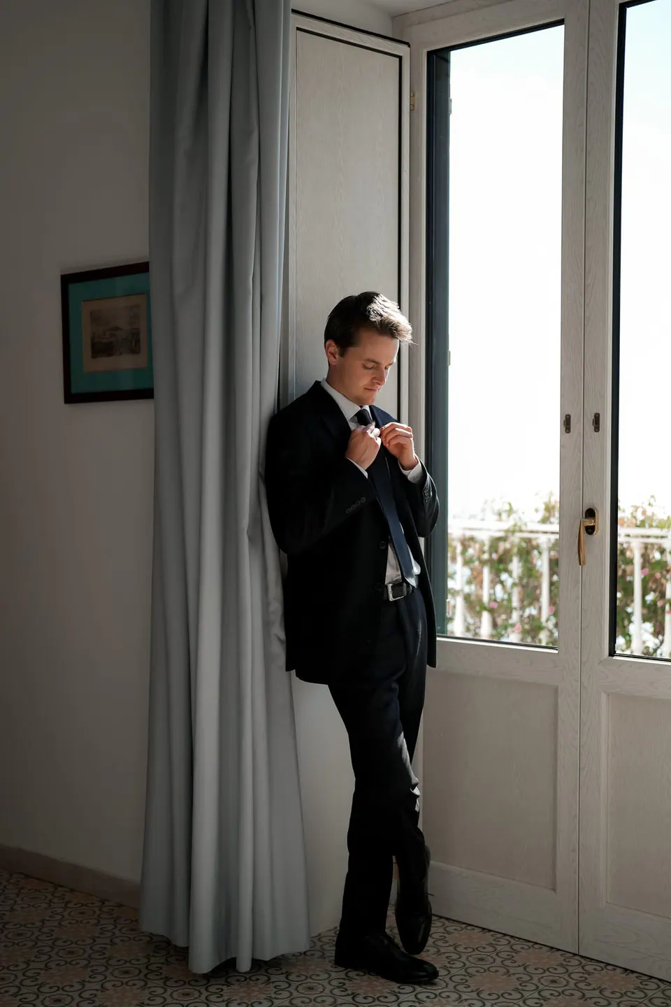 Groom adjusting his tie by the balcony door.