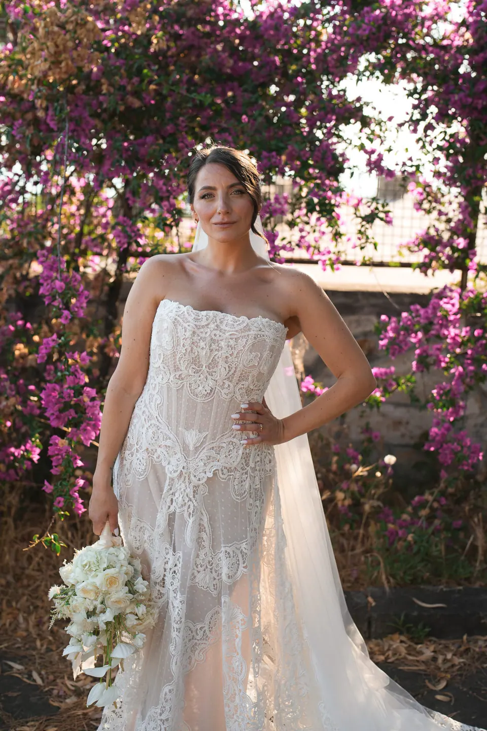 Die Braut im Spitzenkleid vor einer Wand aus Bougainvilleen.