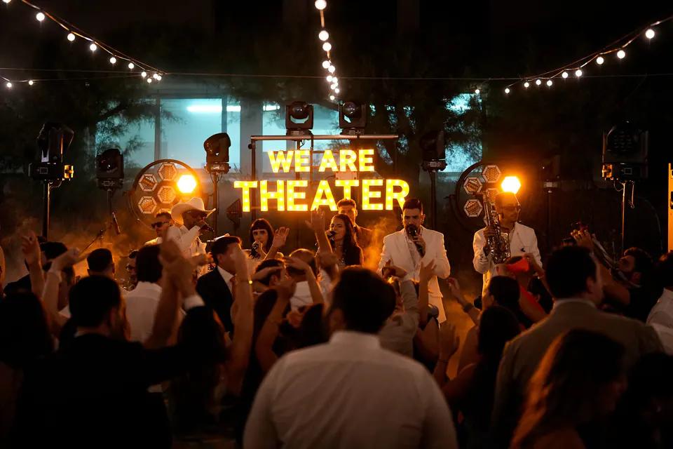 Die Band We Are Theater auf der Bühne, davor tanzende Gäste vor dem Leuchtschild.