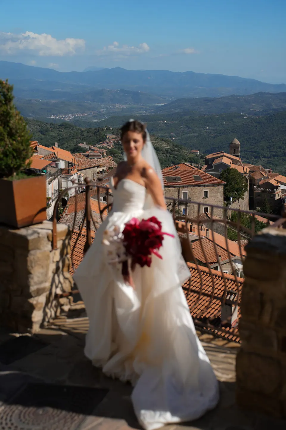 Sposa sorridente in terrazza con il bouquet rosso, tetti e colline sotto.