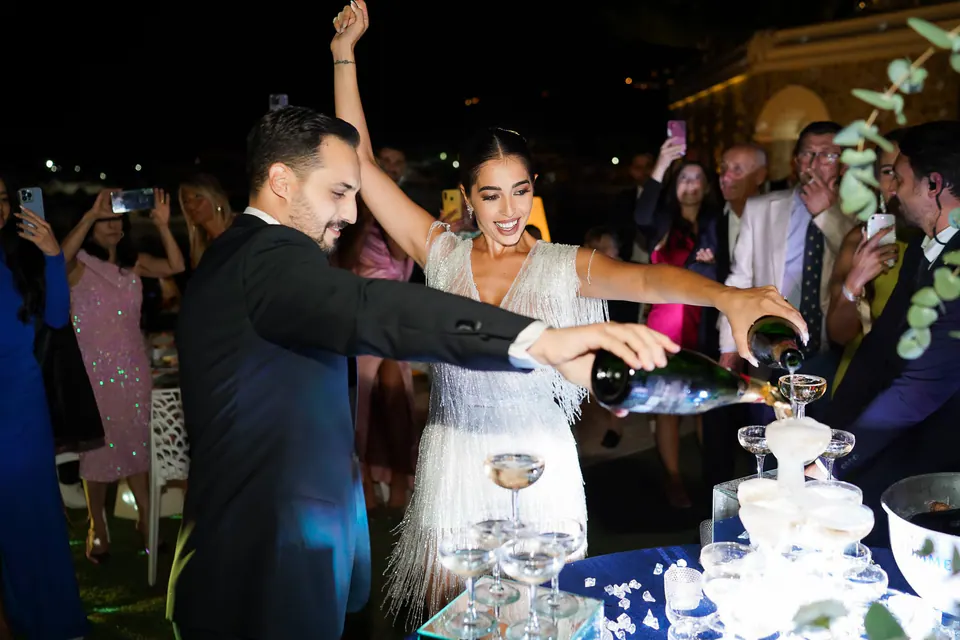 Groom pouring champagne into a tower of glasses as the bride cheers at night.