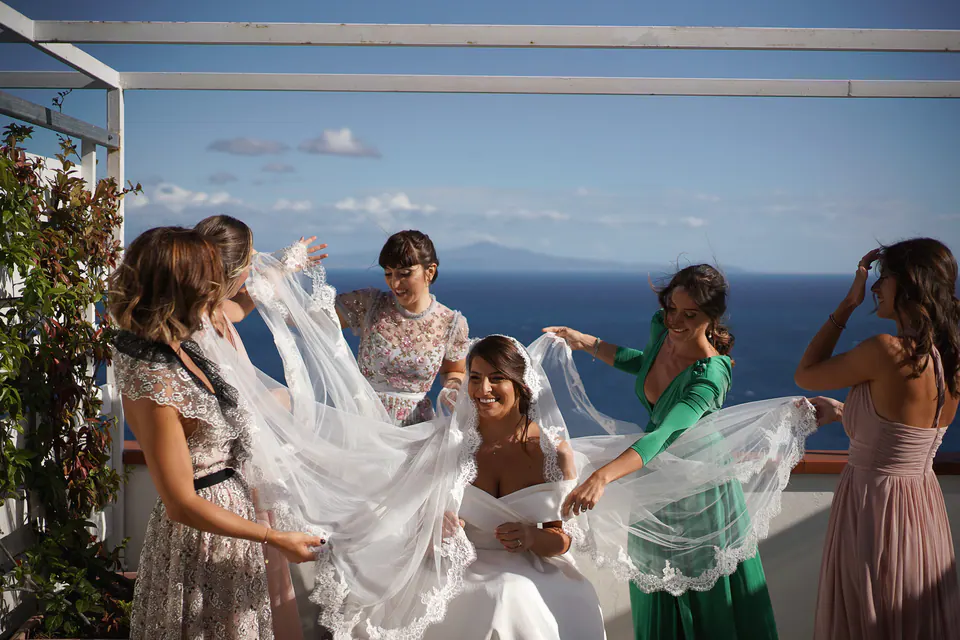 Bridesmaids helping the bride with her veil on the terrace, laughter and sea breeze in the air.