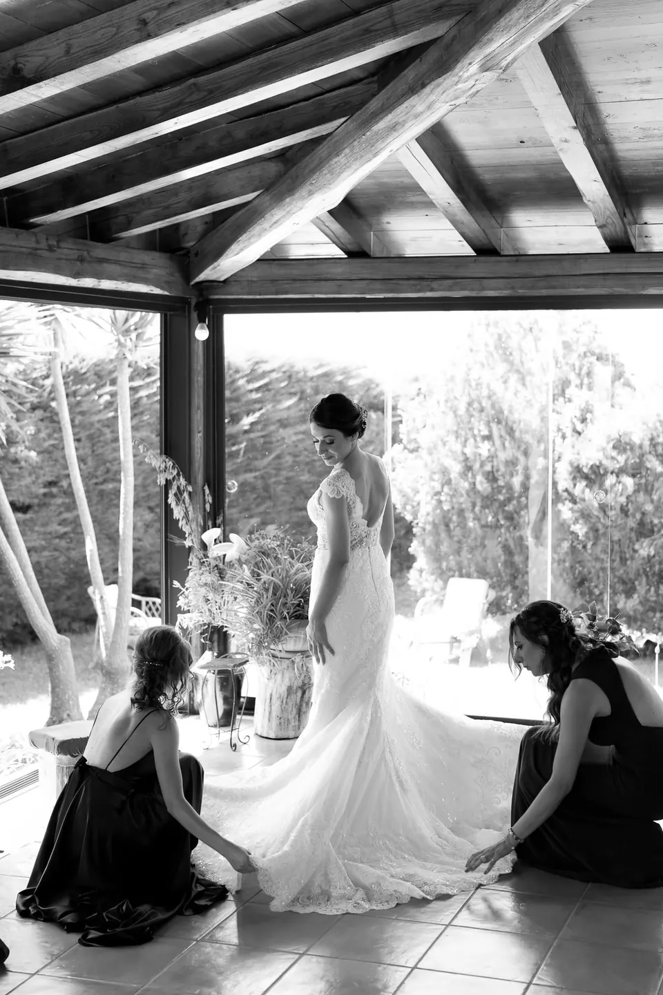 Black and white photo of women arranging the bride's dress under a pergola