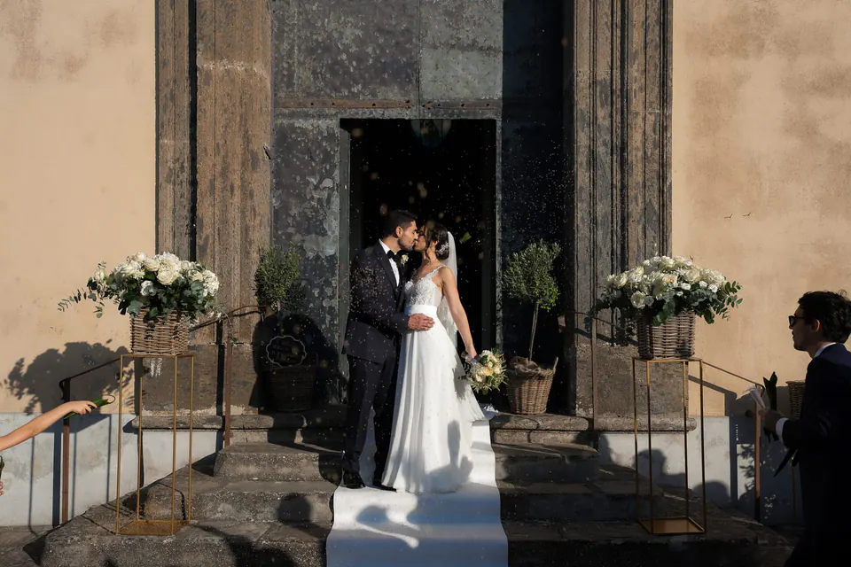 Bride and groom kissing at the church door between arrangements of white flowers.