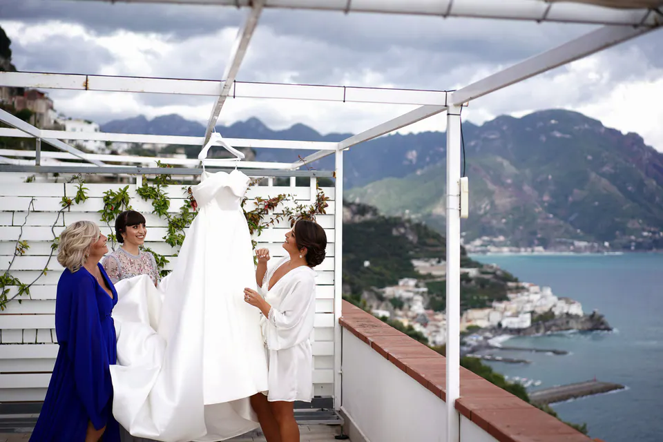 Wedding gown hanging from the terrace pergola as the mother and bridesmaids look on, Amalfi below.