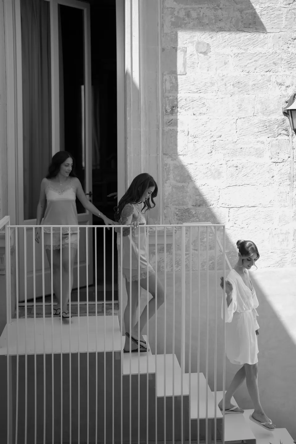 Immagine in bianco e nero della sposa e delle damigelle che scendono una scala bianca al sole.
