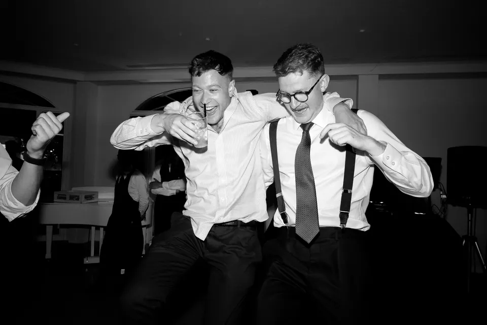 Deux amis en chemise et bretelles dansent bras dessus bras dessous, en noir et blanc.