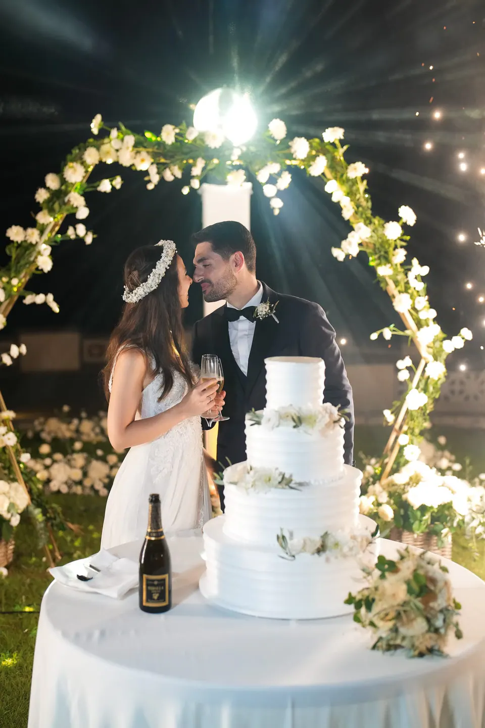 Bride and groom cutting the wedding cake under a floral arch at night.
