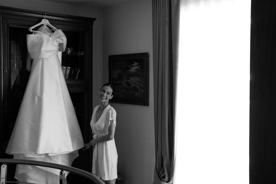 Foto in bianco e nero dell'abito appeso vicino alla finestra mentre la sposa lo guarda.