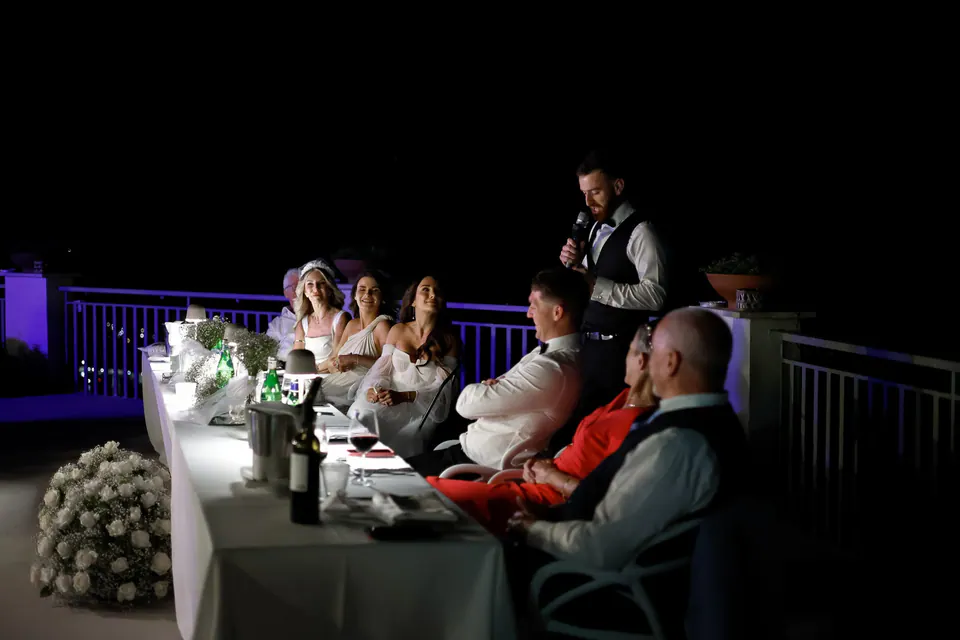 Hochzeitsdinner nachts auf einer Terrasse, die Gäste an der langen Tafel hören einer Rede zu.