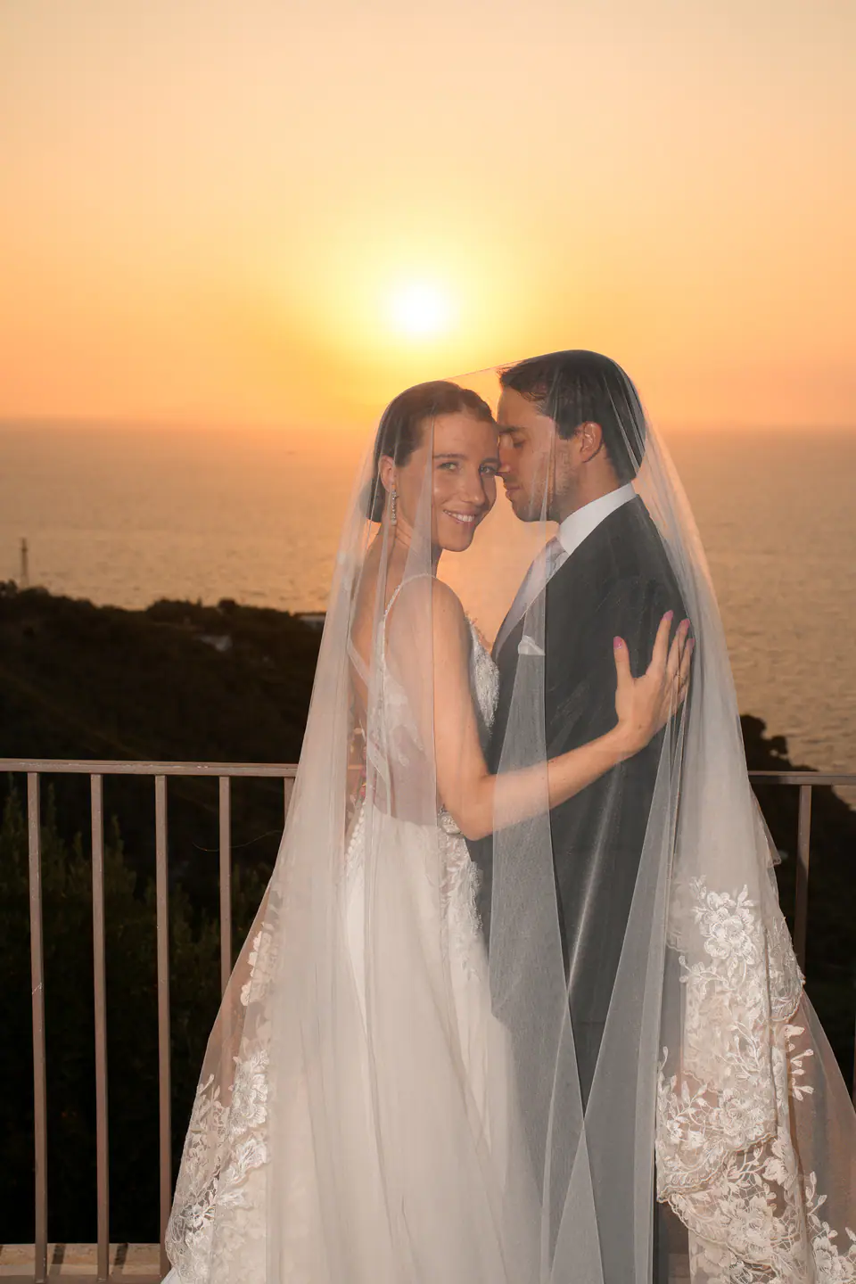 Couple wrapped in the veil at the railing as the sun sets over the sea.