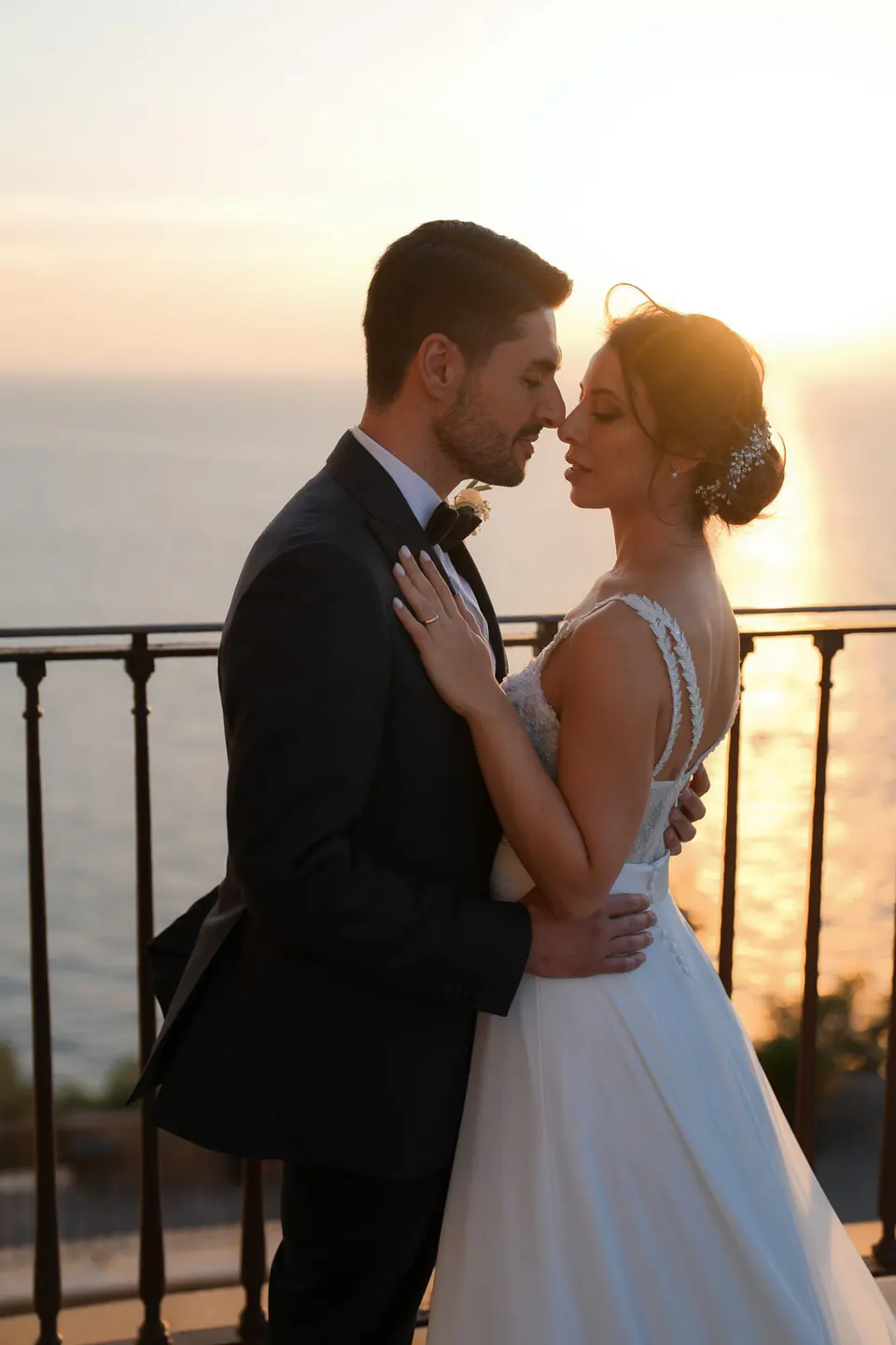 Bride and groom embracing at the railing of Villa Angelina at sunset, the sea behind them.