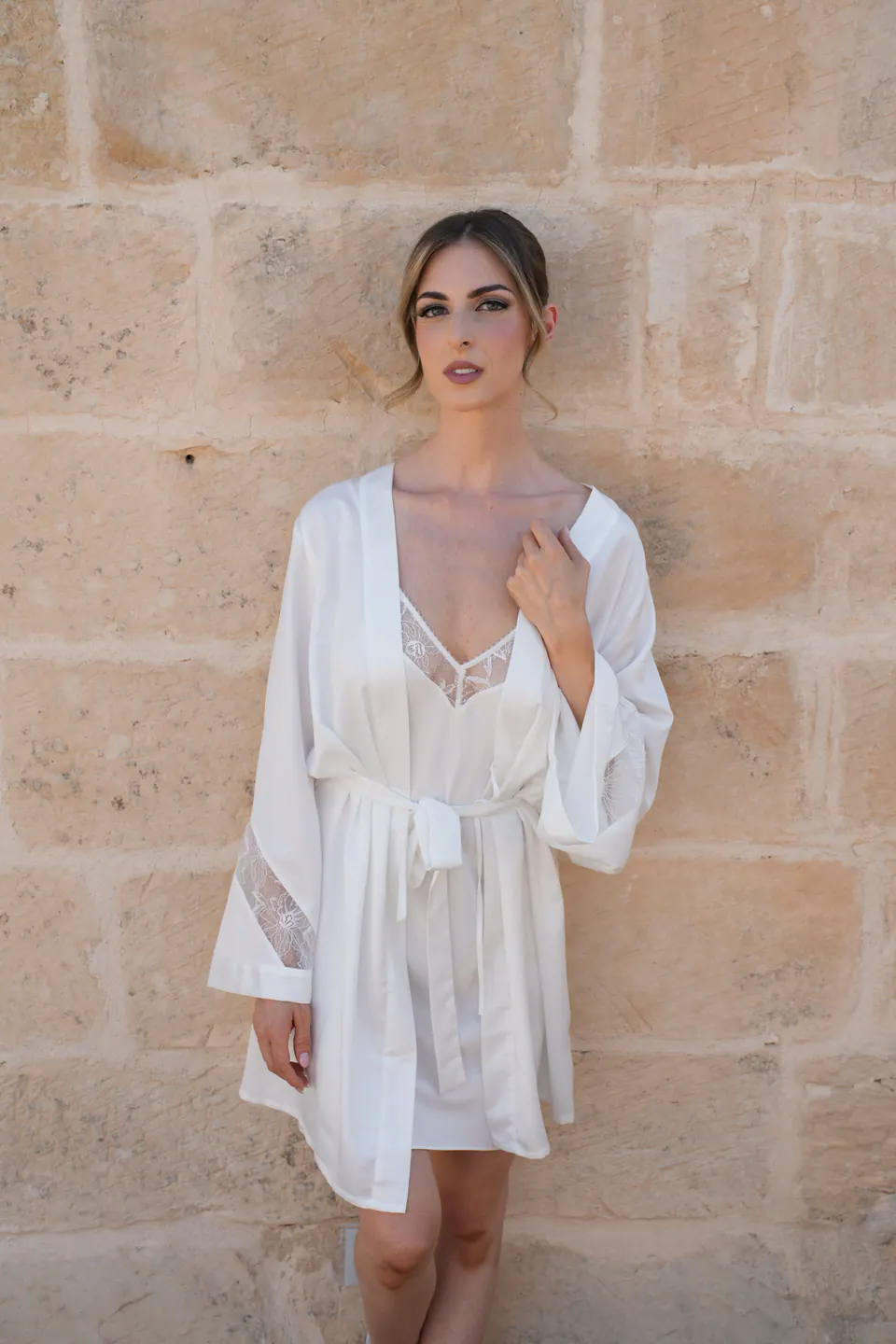 La sposa in vestaglia di seta bianca contro un muro di pietra color miele.