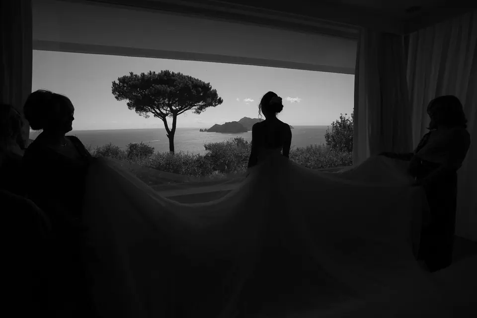 Sagome alla finestra al tramonto, fuori un pino marittimo e il mare.